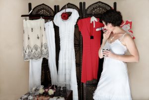 Woman in her 30s-40s standing next to a collection of shoes and other hanging clothes - thinking.